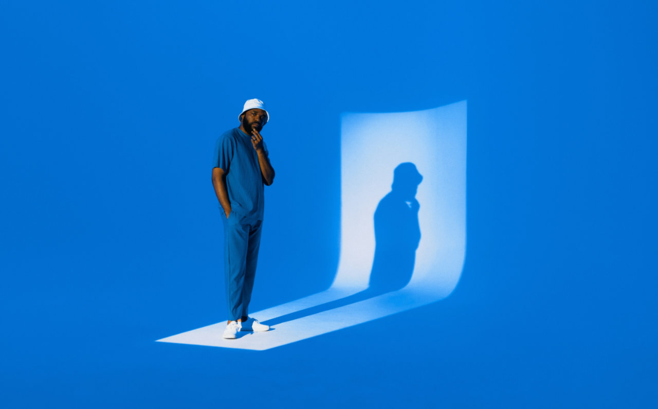 Man wearing a white bucket hat and blue outfit standing on a lit platform with his shadow cast on a curved blue wall.